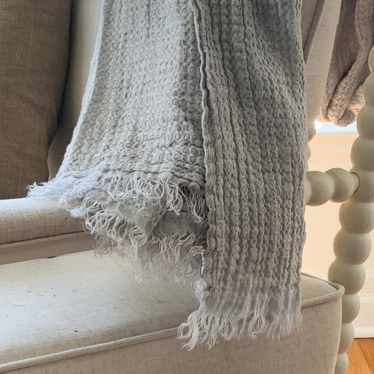 A pale mineral blue textured linen throw blanket with fringed edges draped over a beige upholstered chair in an indoor setting.