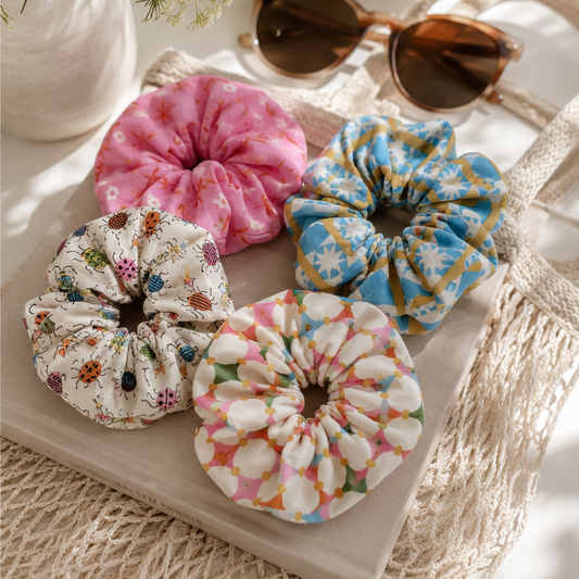 4 fabric hair scrunchies in various floral and polka dot patterns are displayed in a styled flat-lay setting with a woven tray, sunglasses, a glass of water, and decorative vases.