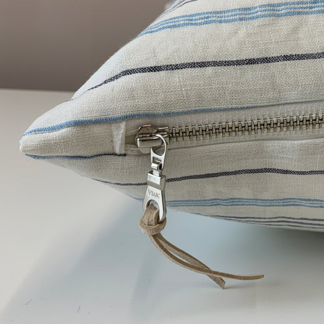 A close-up of a square throw pillow with a white linen fabric body and thin blue and navy stripes, featuring an exposed silver metal zipper pull.