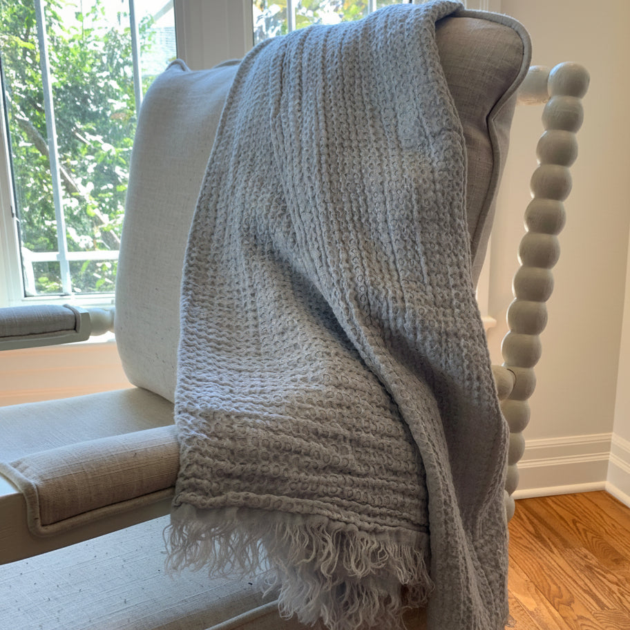 A light mineral blue linen throw blanket with fringe is draped over a beige upholstered armchair in a home interior setting with hardwood floors and a window.