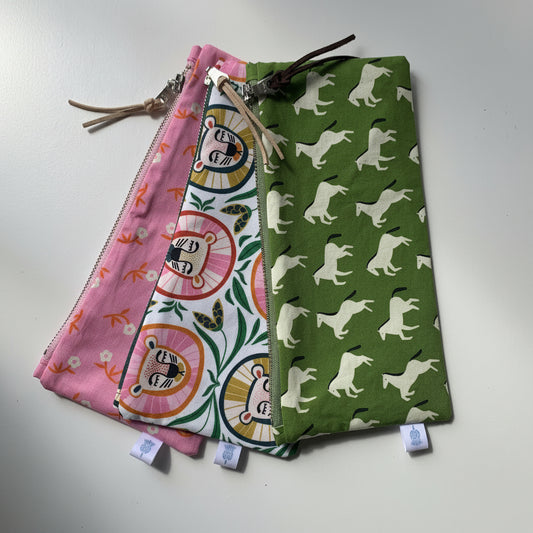 Three fabric zipper pouches with colorful animal and floral prints are arranged overlapping on a white surface. The left pouch is pink with orange and yellow floral details, the middle pouch has a white background with pink and green cartoon lion faces, and the right pouch is olive green with white leaping pony silhouettes.